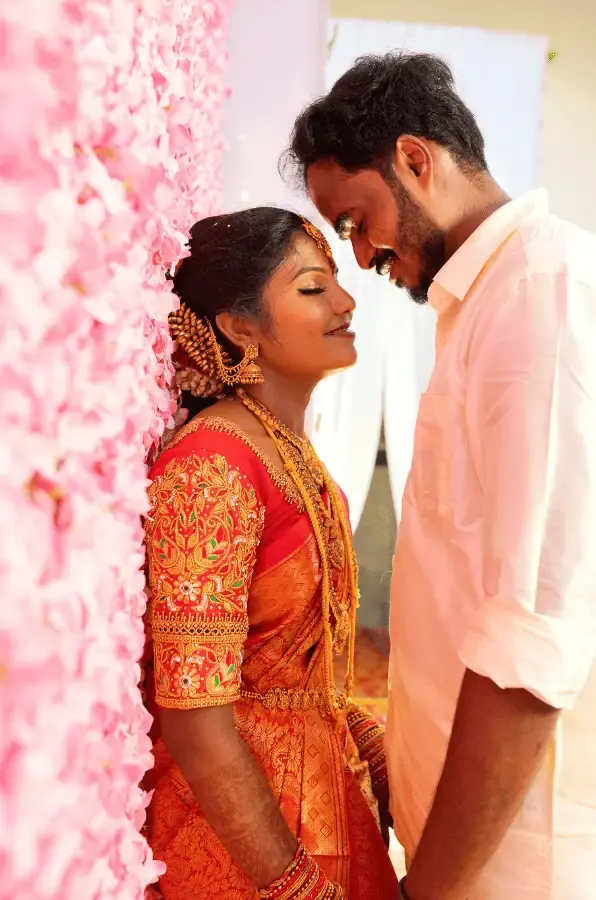 Bride in a red silk saree and groom sharing a close moment against pink flowers